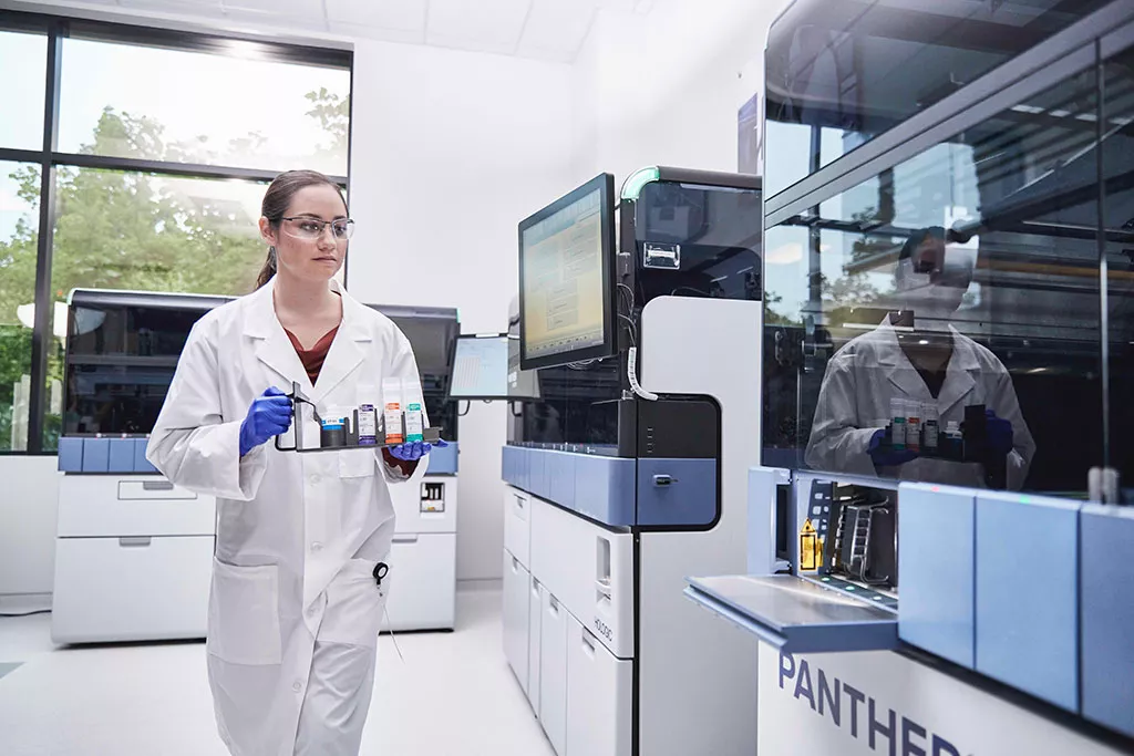 Bioanalytiker, der bærer en bakke med prøvematerialer, der skal sættes i Panther-systemet
